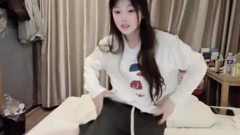 Media: A video shows an Asian woman with long black hair, wearing a white hoodie with a graphic design, and black pants, standing in a bedroom with beige curtains, a cluttered desk, and a bed.