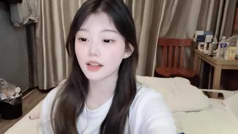 Media: A video of an Asian woman with long, straight black hair, fair skin, and light makeup, wearing a white shirt, sitting on a bed in a cluttered room with beige curtains and wooden furniture.