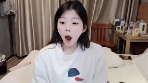 Media: Video of a young Asian woman with long black hair, wearing a white hoodie, mouth open in an expression of surprise or shock, in a cluttered bedroom with beige curtains and a wooden chair.