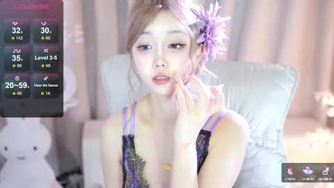 Media: Video of a young East Asian woman with fair skin, wearing a purple lace bra, sitting on a white couch. She has long, brown hair adorned with a large, pink flower. Background includes a teddy bear and soft lighting.