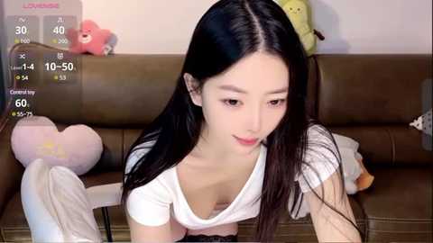 Media: Video of a young Asian woman with long black hair, wearing a white top, sitting on a brown couch with stuffed toys.