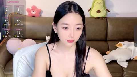 Media: A video of an East Asian woman with long black hair, wearing a black spaghetti-strap top, sitting on a brown leather couch in a room with stuffed toys and a white blanket.