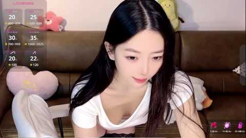 Media: A video of an East Asian woman with long black hair, light skin, and a slender physique, wearing a white top, sitting on a brown leather couch in a cozy room.