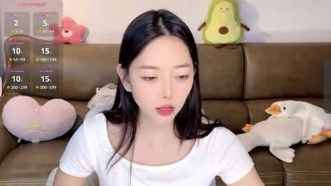 Media: A video of a young East Asian woman with long black hair, wearing a white t-shirt, sitting on a brown leather sofa. The background features plush toys, including a green duck and a pink bear.