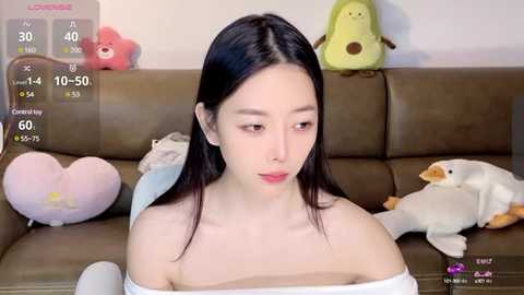 Media: A video of a young Asian woman with long black hair, sitting on a brown leather couch, surrounded by plush toys. She wears a white off-shoulder top.