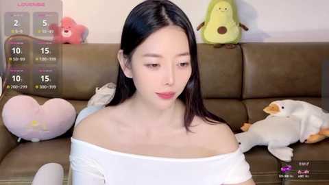 Media: A video of an East Asian woman with long black hair, fair skin, and a white off-shoulder top, sitting on a brown leather couch. She's surrounded by plush toys, including a yellow duck and a heart-shaped pillow.