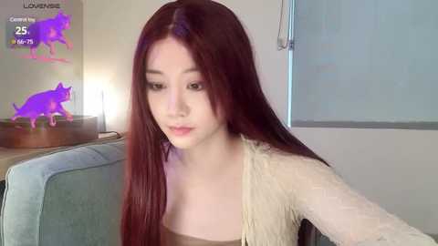 Media: Video of a young Asian woman with long, straight, auburn hair, pale skin, wearing a light-colored top, seated in a light blue armchair. Background includes a purple cat figurine on a wooden table and a window with closed blinds.