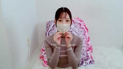 Media: A video of an East Asian woman with dark hair and fair skin, wearing a beige striped sweater and a white surgical mask, seated on a white bed with a pink and white patterned pillow behind her.