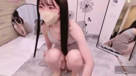 Media: Video of a pale-skinned, petite Asian woman with long black hair and bangs, squatting in a light-colored room. She wears a white face mask, revealing her flushed cheeks. A large mirror reflects her image, and the background features minimalist decor.