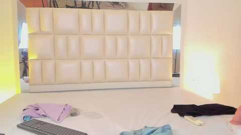 Media: Video of a minimalist room with a white, cushioned wall panel, yellow light on the left, scattered clothes on the floor, and a remote control.