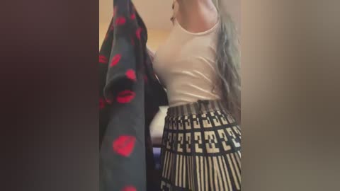 Media: Video of a woman with fair skin, wearing a white tank top and high-waisted, patterned pants, holding a red polka-dotted jacket, in a dimly lit room.