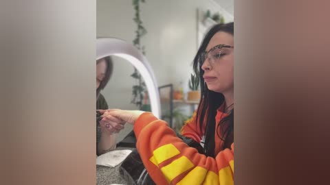 Media: Video of a young woman with long black hair, wearing glasses and an orange and yellow striped sweater, seated at a desk in a modern, minimalist room with a large round mirror and plants.