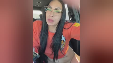 Media: Video of a Latina woman with long black hair, wearing glasses and a cropped red shirt, making a playful face while seated in a car.