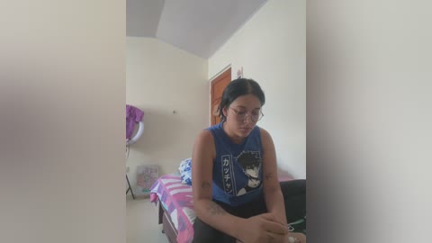 Media: A video of a young woman with medium skin tone, dark hair, and glasses, wearing a blue tank top with a graphic design, seated in a sparsely furnished room with a pink bed and a chair.