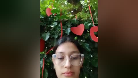 Media: Video of a young woman with light brown skin, dark hair, and clear glasses, standing in front of green foliage adorned with red hearts, creating a romantic, whimsical atmosphere.