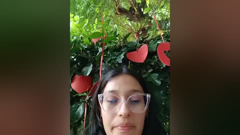 Media: Video of a young woman with long black hair, wearing clear-framed glasses, lying on a bed of green leaves. Her face is serene, surrounded by red heart-shaped decorations and lush foliage.