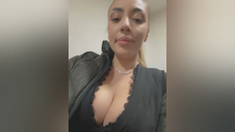 Media: Video of a young, light-skinned woman with a slim physique, wearing a black lace bra and unzipped black leather jacket, with a close-up focus on her cleavage and face, in a neutral indoor setting.