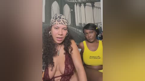 Media: Video of two women in a selfie; one with curly hair, leopard-print headband, and low-cut red lace top, the other with straight hair, yellow crop top, and black hairband. Background features a black-and-white mural of a bridge.