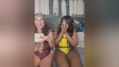 Media: Video of two women posing in lingerie: one with a leopard print headband, maroon lace bra, and panties, the other with yellow lingerie. Background features a large, black-and-white mural of the Brooklyn Bridge.