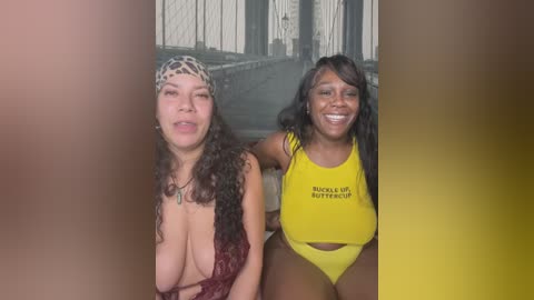 Media: Video of two women; one with long curly hair, large breasts, and a leopard-print headband, wearing a low-cut top; the other with long straight hair, medium breasts, wearing a yellow crop top. Background shows a blurred urban scene.