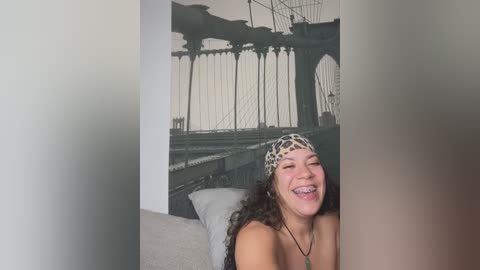 Media: Video of a joyful, smiling woman with curly hair and a leopard print headband, wearing a black top, seated in a room with a large, monochrome Brooklyn Bridge mural on the wall.