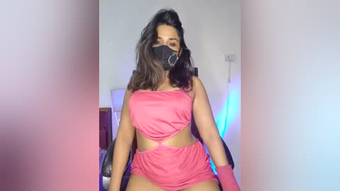 Media: A video of a Latina woman with medium skin tone and long, wavy brown hair. She wears a black face mask, a pink crop top, and matching shorts, sitting in a dimly lit room with neon blue and pink lighting.