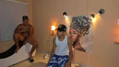 Media: Video of a beige-walled room with a man sitting shirtless on a bed, a boy in a tank top and blue shorts, and a large African art print.