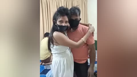 Media: Video of two South Asian women in white dresses and black masks, standing close together, with beige curtains in the background.