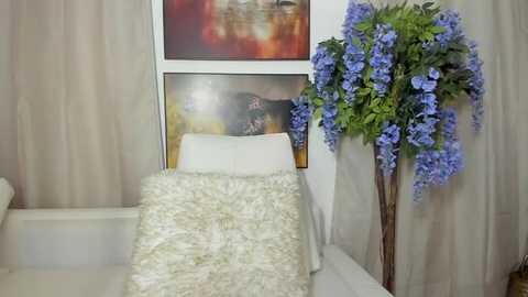 Media: A video of a cozy room with a white chair and fluffy white pillow, a large vase of cascading blue flowers, and abstract artwork on beige walls.