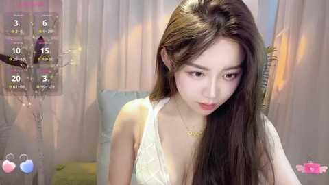 Media: Video of an East Asian woman with long black hair and fair skin, wearing a white lace top, sitting in a softly lit room with a digital clock and pink flowers.