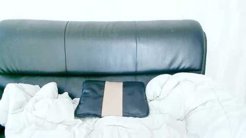 Media: A video of a black leather couch with a beige pillow and a black cushion, covered in white, crumpled blankets, creating a cozy, messy bedroom scene.