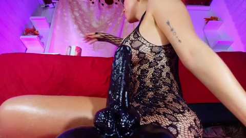 Media: Video of a light-skinned woman in a black lace dress, seated on a red couch, wearing fishnet stockings, holding a large, dark, textured dildo. Background includes a white wall with decorative items.