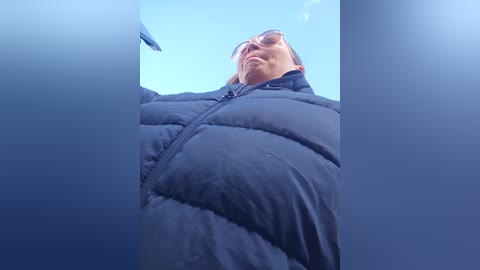 Media: A video shows a man in a black puffer jacket, wearing sunglasses and looking up, against a clear blue sky. The image is slightly blurred, emphasizing the sky's depth.