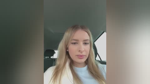 Media: A video of a blonde Caucasian woman with long hair, wearing a light-colored jacket, sitting in a car with a dark interior, looking contemplative.