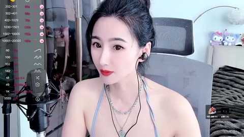 Media: Video of an East Asian woman with fair skin, dark hair tied back, wearing red lipstick, a light gray halter top, and multiple necklaces. Background includes a bed with a gray blanket and stuffed animals.