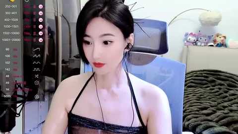 Media: A video of a young Asian woman with fair skin, black hair in a bun, and red lipstick, wearing a black lace top, sitting in a bedroom with a plush toy, a bed, and a video screen showing live streaming stats.