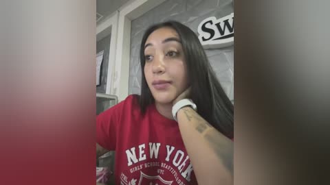 Media: Video of a young woman with medium skin tone, long black hair, and a red \"New York\" t-shirt. She's leaning against a wall with a \"SW\" sign and white brick background.