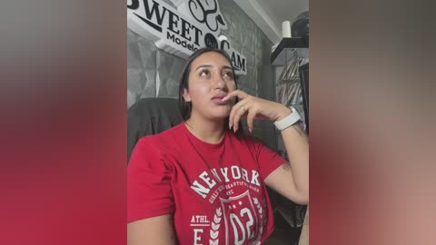 Media: Video of a young Latina woman with medium skin tone, sitting in a black chair, wearing a red t-shirt with white text, and resting her chin on her hand. Background features a gray wall with decorative text and a shelf.