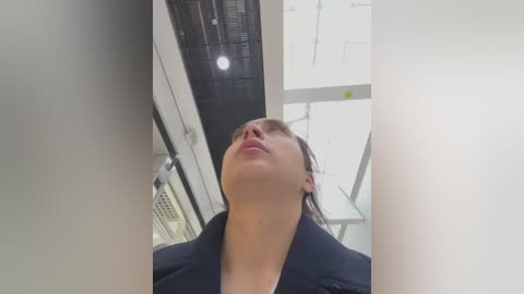 Media: Video of a woman with short brown hair, wearing a dark blazer, looking up in a modern, well-lit room with white walls and metallic ceiling elements.