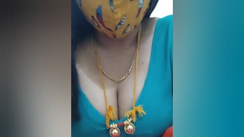 Media: Video of a woman with fair skin, wearing a blue dress with a low neckline, gold necklace with two dangling yellow tassels, and a yellow face mask with cartoon characters, standing in a blue-tinted room.