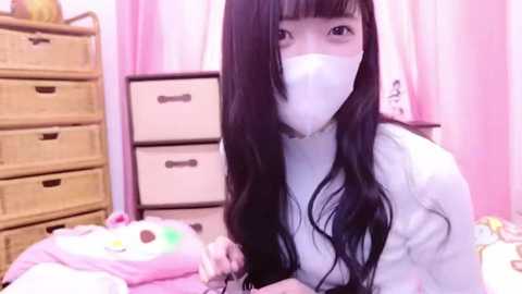 Media: A video of an Asian woman with long black hair, wearing a white mask and a white sweater, holding a stuffed animal, standing in a pink room with white drawers and a window.