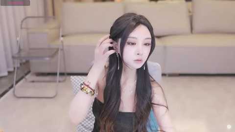 Media: A young Asian woman with long black hair, fair skin, and wearing a black top, adjusts her hair while seated on a beige sofa in a modern living room with a glass coffee table and white curtains.