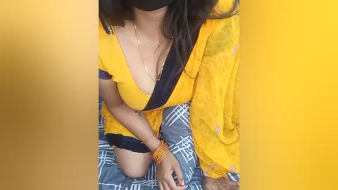 Media: Video of a South Asian woman with medium brown skin and long dark hair, wearing a yellow sari with a black trim, seated on a blue-and-white plaid bedspread, hands bound with yellow bracelets.