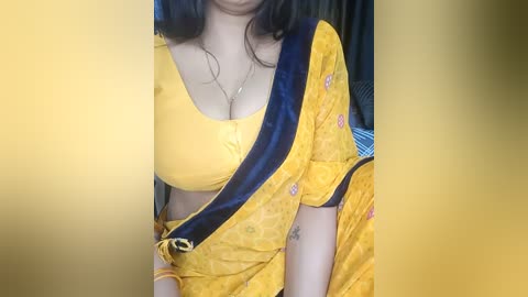 Media: Video of a South Asian woman wearing a yellow sari with a blue border, showcasing her medium-sized breasts in a low-cut blouse. She has medium skin tone and wears gold jewelry.