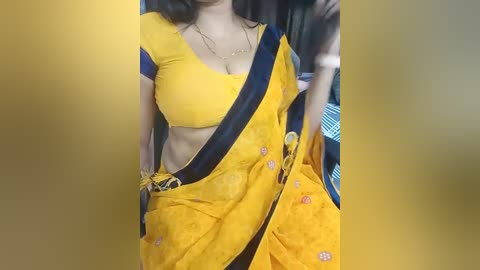 Media: Video of a South Asian woman in a bright yellow sari with blue and pink floral embroidery, wearing a matching blouse, with a blue shoulder strap and a gold belt.