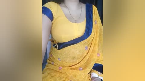 Media: A video of a light-skinned woman in a bright yellow saree with blue borders and floral patterns, wearing a gold necklace. Her medium-sized breasts are visible through the thin fabric. The background is blurred.