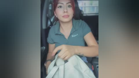 Media: Video of a young woman with medium skin tone, red hair, and a small tattoo on her wrist, wearing a gray polo shirt, holding a white bag in a dimly lit car interior.