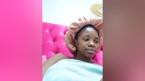 Media: Video of a young Black woman with short hair, lying on a pink tufted leather couch, covered with a light blue towel, against a white wall with a golden sunburst decoration.