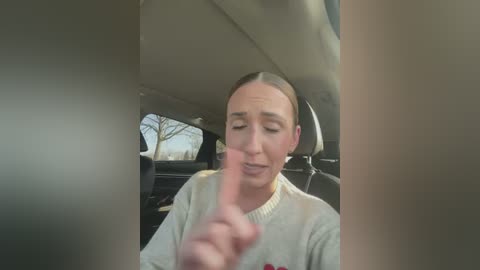 Media: A video of a woman with short brown hair, wearing a light-colored sweater, sitting in a car, holding a finger up, with a blurry tree visible through the window.