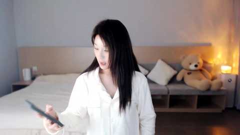 Media: Video of a young woman with long black hair, wearing a white robe, holding a tablet, standing in a minimalist bedroom with a beige headboard, white bedding, and a plush teddy bear.